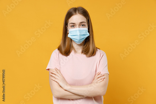 Young woman 20s in basic pastel pink t-shirt, blank print design in sterile face mask to safe from coronavirus covid-19, pandemic quarantine hold hands crossed folded isolated on yellow background