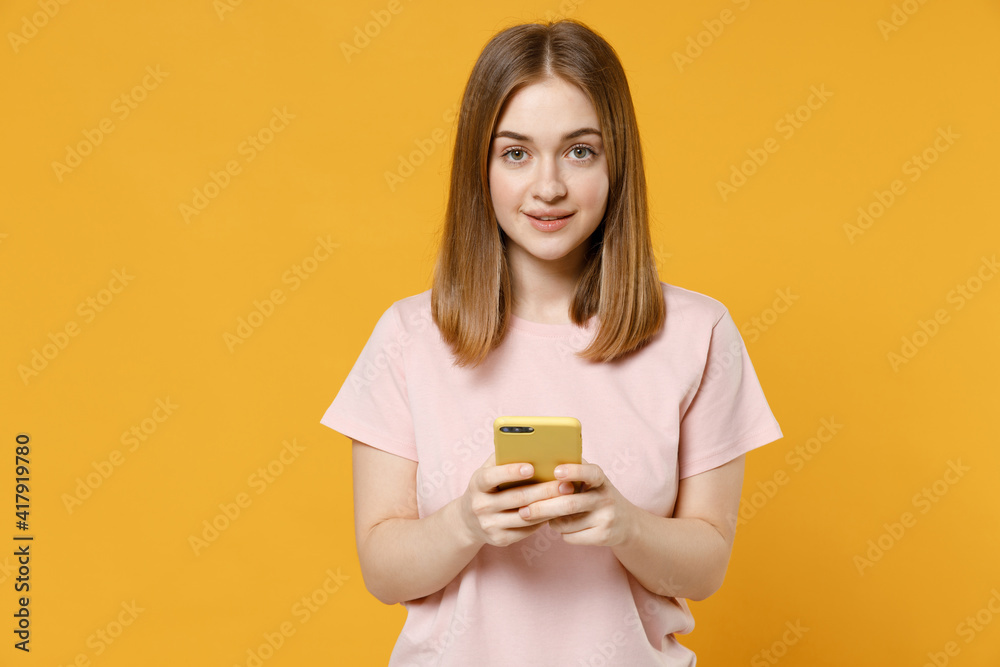 Young caucasian woman 20s with nude make up wearing casual basic pastel pink t-shirt hold mobile cell phone surfing browsing internet looking camera isolated on yellow background studio portrait Young caucasian woman 20s with nude make up wearing casual basic pastel pink t-shirt hold mobile cell phone surfing browsing internet looking camera isolated on yellow background studio portrait