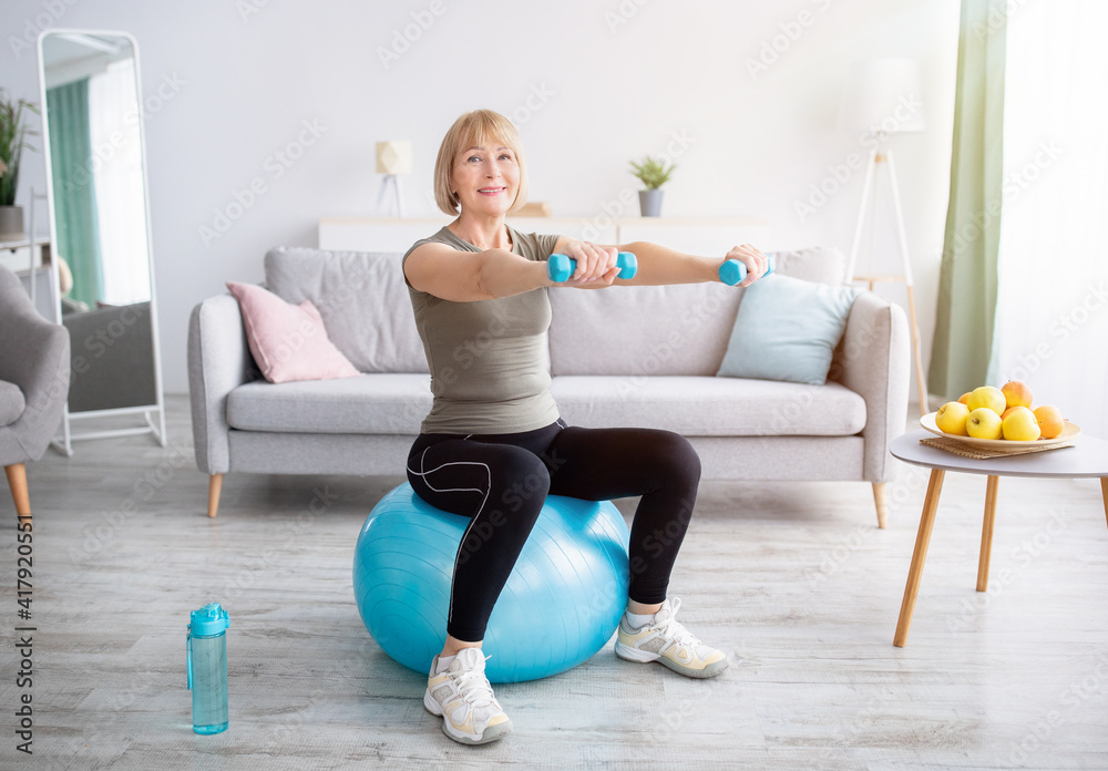Fototapeta premium Lovely mature woman sitting on fitness ball, working out with dumbbells in her home gym, empty space. Active lifesyle