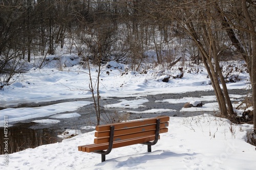 The empty bench by the creek in the snow on a winter day.