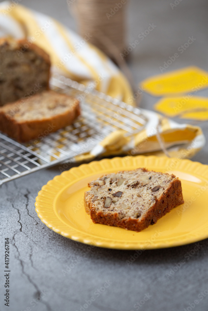 A Slice of Banana Nut Bread on a Yellow Plate with Loaves on Cooling Rack in Background; Twine and Banan Nut Bread Tags in Background; Gray Background