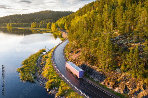 Aerial view of semi truck with cargo trailer on road curve at lake shore with green pine forest. Transportation background. Beautiful nature landscape at sunset in Republic of Karelia, Russia