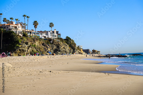 Wallpaper Mural a gorgeous shot of people relaxing on a sandy beach near a lush green hillside with homes and palm trees with ocean water and blue sky  at West Street Beach in Laguna Beach California