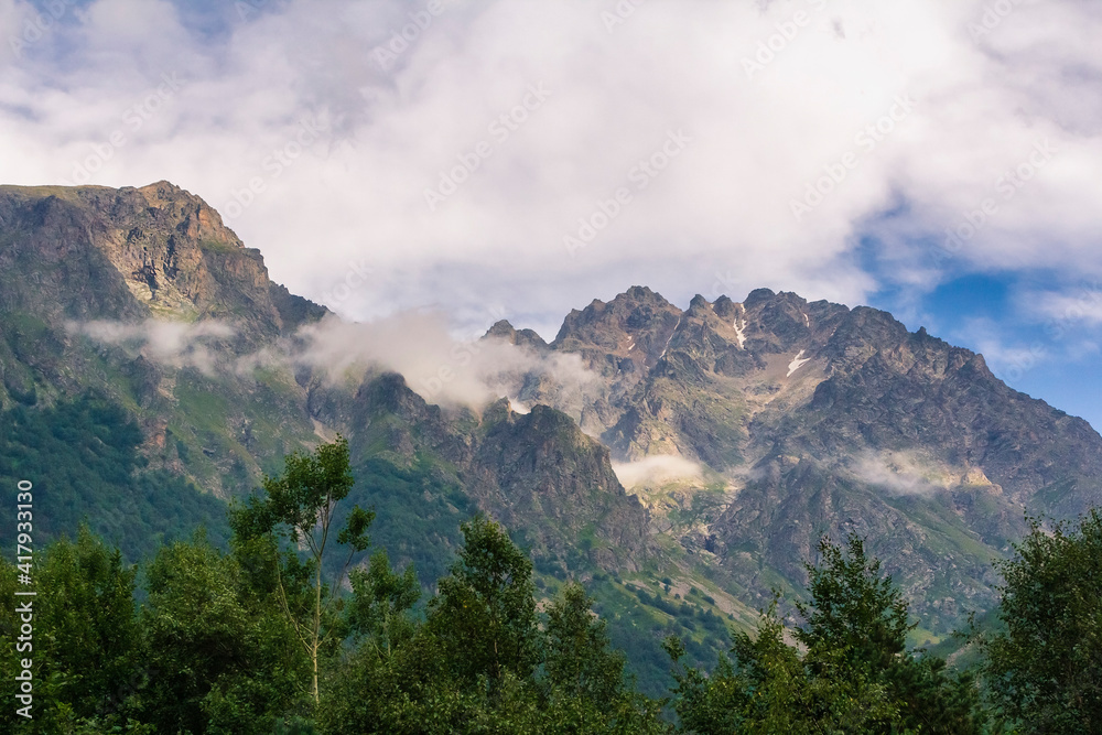 High mountains of the Caucasus with a beautiful view. Green vegetation and shrubs on sharp rocks above a blue sky. Magnificent mesmerizing landscape.