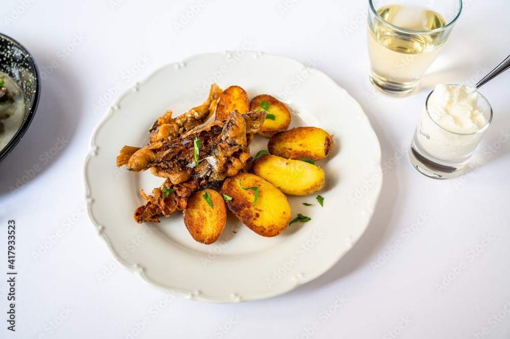 Fried sardines with potato on white plate, garlic yogurt in glass, wine and part of pan Fried sardines with potato on white plate, garlic yogurt in glass, wine and part of pan