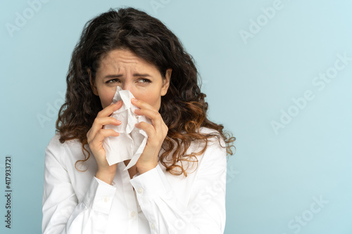 Studio portrait of young office employee woman in white blouse with tissue blowing nose, isolated on blue background, copy space. Rhinitis, seasonal allergy, symptoms of flu virus concept.