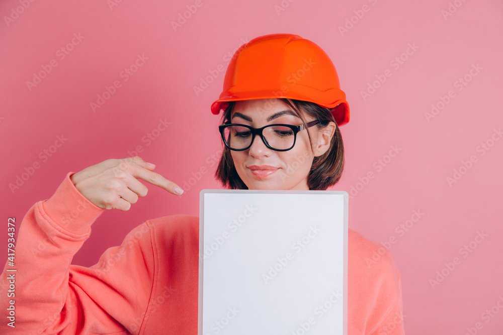 Smiling woman worker builder hold white sign board blank against pink background. Building helmet. Point index finger. Smiling woman worker builder hold white sign board blank against pink background. Building helmet. Point index finger.