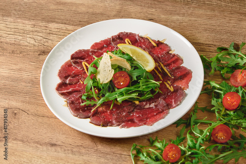 Carpaccio sliced meat with salad, tomatoes and lemon on a white plate on a wooden table.