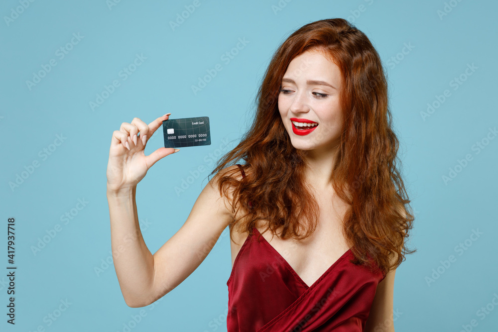 Young caucasian smiling cute happy rich successful ginger readhead curly woman 20s wear red party evening dress gown holding credit bank card isolated on pastel blue color background studio portrait.