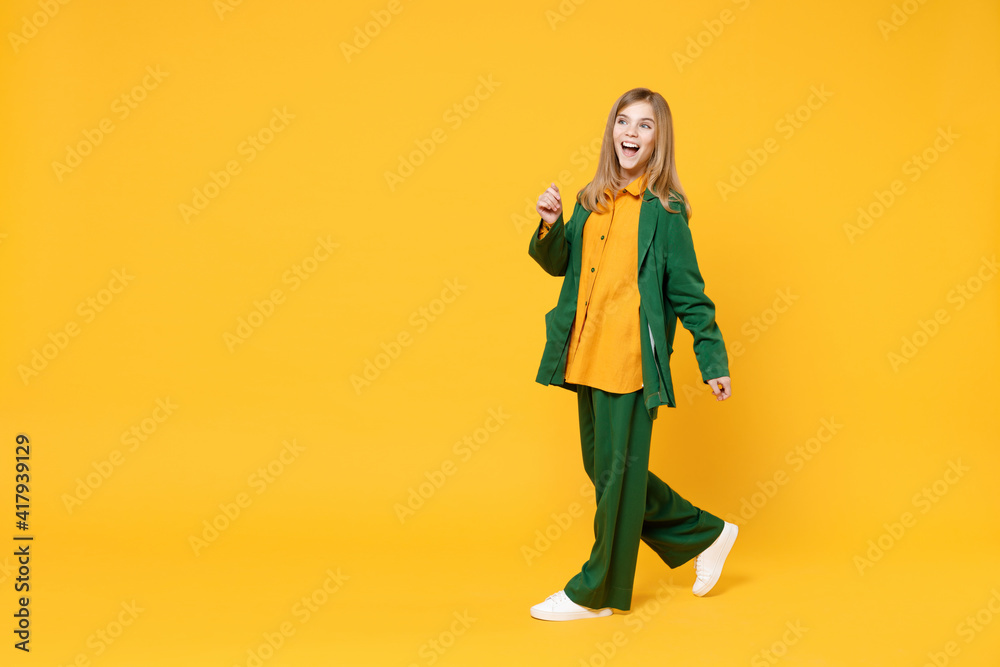 Full length little blonde kid happy girl 12-13 years old wearing casual clothes walking smiling look aside isolated on yellow orange background children studio portrait. Childhood lifestyle concept Full length little blonde kid happy girl 12-13 years old wearing casual clothes walking smiling look aside isolated on yellow orange background children studio portrait. Childhood lifestyle concept