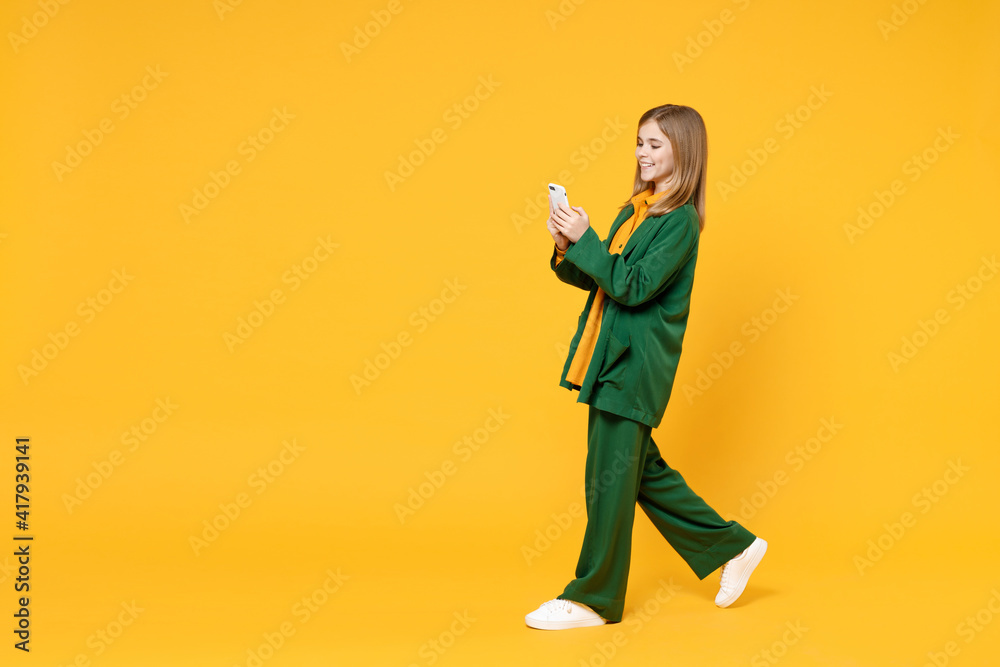 Full length side profile view of little blonde cute kid girl 12-13 years old wearing casual clothes hold mobile cell phone walk go isolated on yellow background children Childhood lifestyle concept. Full length side profile view of little blonde cute kid girl 12-13 years old wearing casual clothes hold mobile cell phone walk go isolated on yellow background children Childhood lifestyle concept.