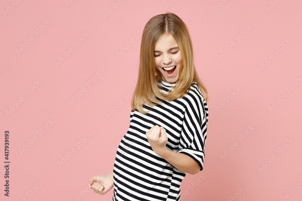 Little blonde excited kid girl 12-13 years old wearing striped oversized t-shirt do winner gesture clench fist isolated on pastel pink background children studio portrait. Childhood lifestyle concept. Little blonde excited kid girl 12-13 years old wearing striped oversized t-shirt do winner gesture clench fist isolated on pastel pink background children studio portrait. Childhood lifestyle concept.