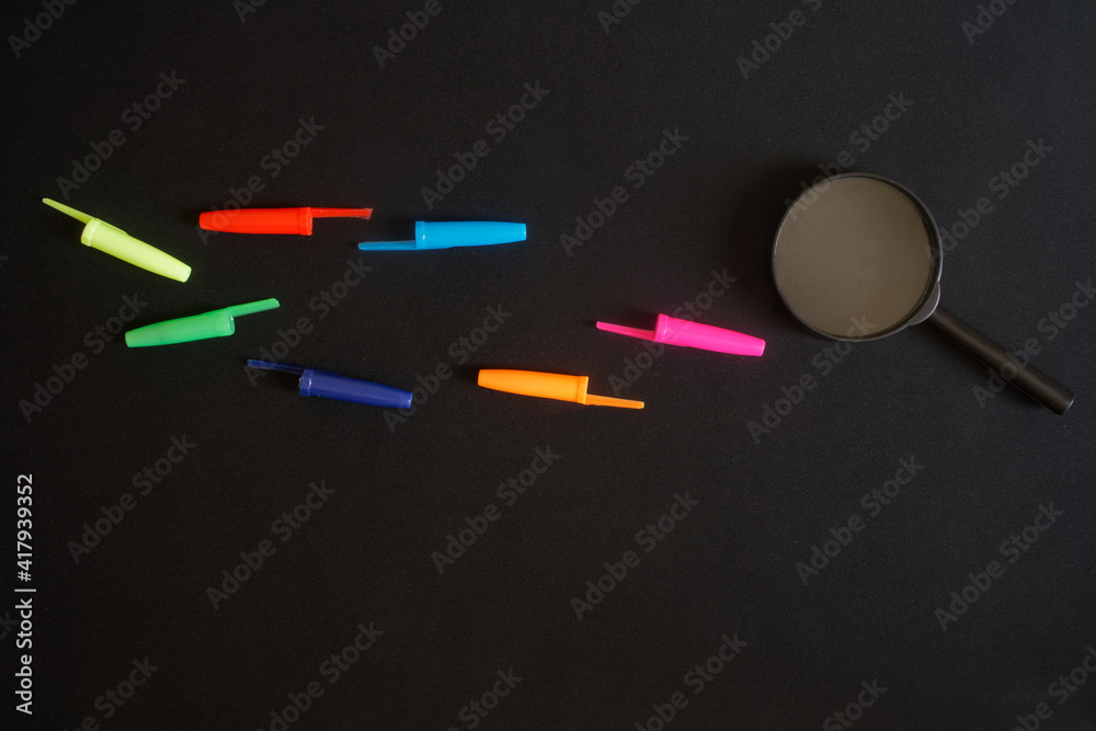 The magnifying glass lies next to the multi-colored pen caps. Search and investigation concept and metaphor. Search for ballpoint pen on traces The magnifying glass lies next to the multi-colored pen caps. Search and investigation concept and metaphor. Search for ballpoint pen on traces