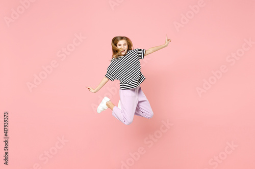 Full length little blonde kid girl 12-13 years old in striped oversized t-shirt jump high with outstretched hands look camera isolated on pastel pink background children. Childhood lifestyle concept.