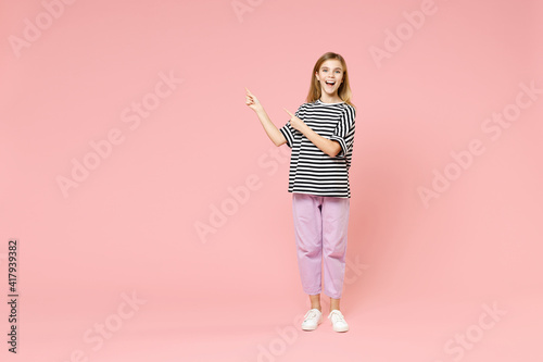 Full length little blonde kid girl 12-13 years old in striped oversized t-shirt point index finger aside on workspace area isolated on pink background children portrait. Childhood lifestyle concept.
