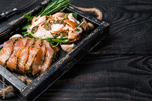 Toasts with hot and cold smoked salmon, arugula in a wooden tray with herbs. Black wooden background. Top view. Copy space