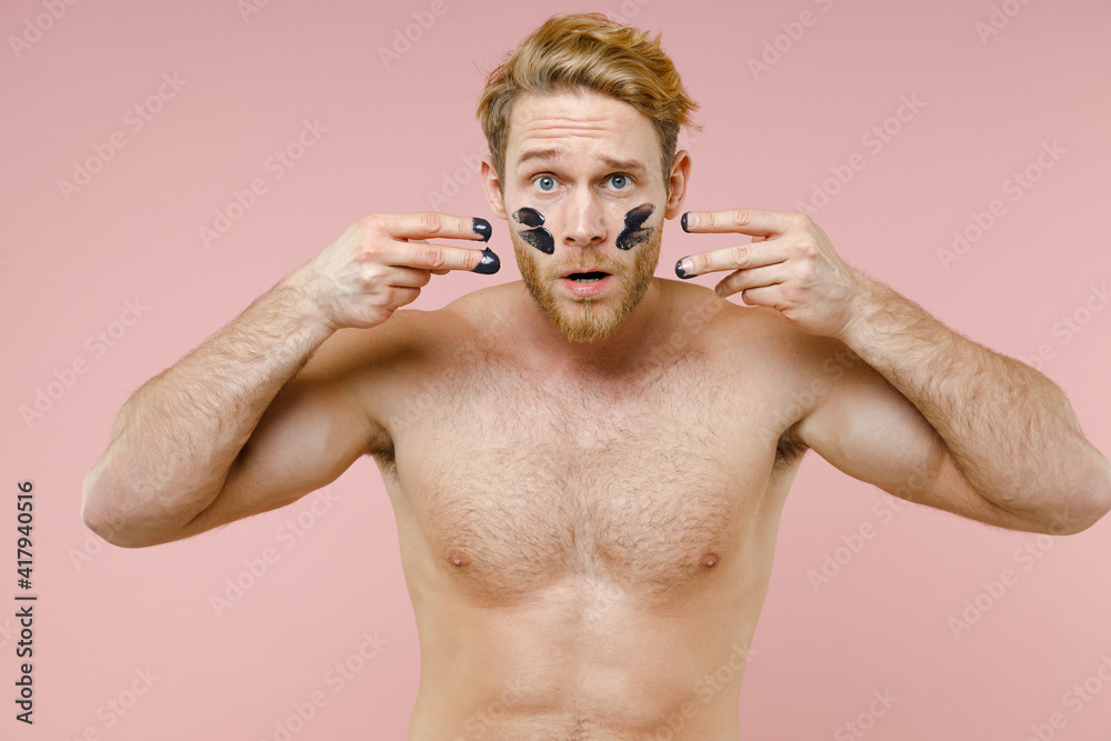 Fototapeta premium Shocked bearded naked young man 20s years old perfect skin smear clay facial mask on cheek isolated on pastel pink color background studio portrait. Skin care healthcare cosmetic procedures concept.
