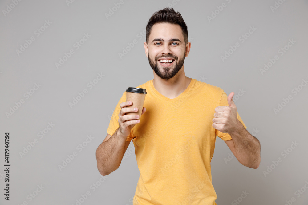 Young caucasian smiling bearded attractive freelancer man 20s wearing casual yellow basic t-shirt drinking hold paper cup of coffee show thumb up gesture isolated on grey background studio portrait.