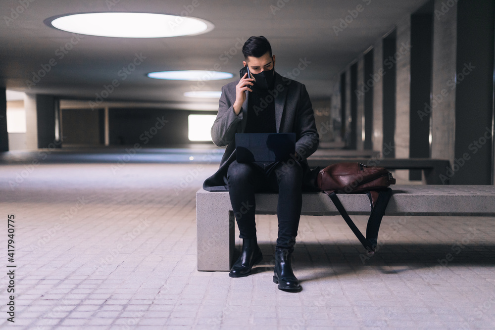 Masked businessman calling with his cell phone while sitting on a bench with his laptop. Masked businessman calling with his cell phone while sitting on a bench with his laptop.
