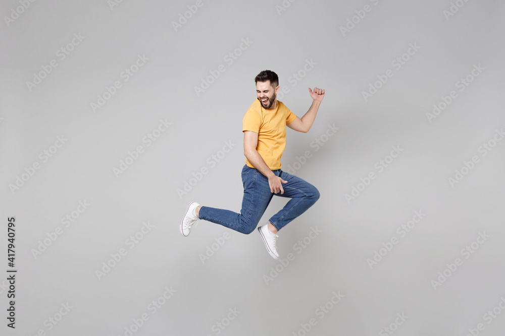 Full length of young bearded student smiling happy overjoyed man 20s in yellow basic t-shirt jump high playing guitar gesture flying outstretched hands isolated on grey background studio portrait Full length of young bearded student smiling happy overjoyed man 20s in yellow basic t-shirt jump high playing guitar gesture flying outstretched hands isolated on grey background studio portrait