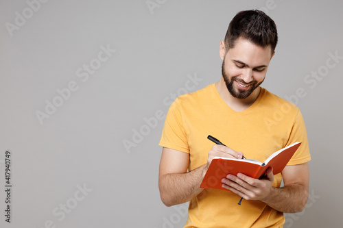 Photography Young caucasian smiling bearded attractive active happy student smart man 20s wearing casual yellow basic t-shirt writing down in exercise book diary isolated on grey color background studio portrait