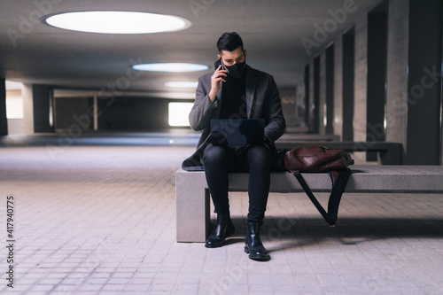 Masked businessman calling with his cell phone while sitting on a bench with his laptop.