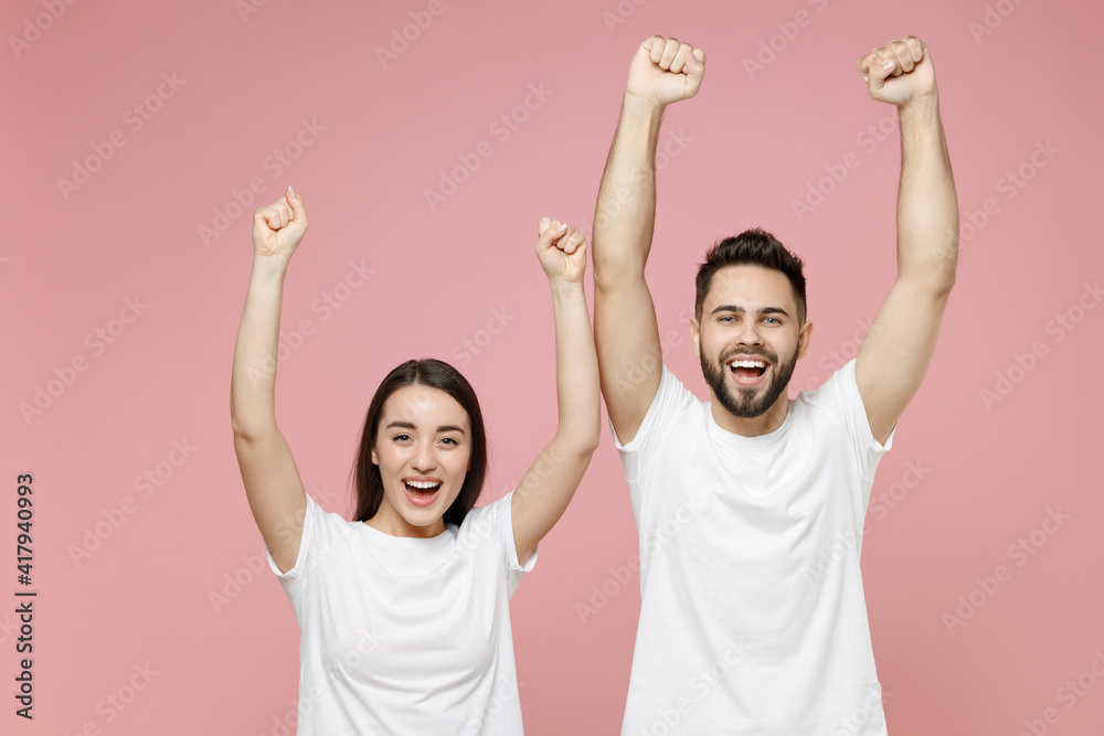 Young cheerful couple two friends man woman in white basic blank print design t-shirt doing winner gesture celebrating clenching fists say yes isolated on pastel pink color background studio portrait Young cheerful couple two friends man woman in white basic blank print design t-shirt doing winner gesture celebrating clenching fists say yes isolated on pastel pink color background studio portrait