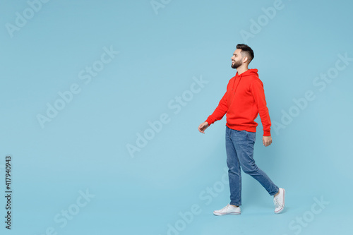 Full length side profile view of young caucasian smiling bearded handsome student man 20s in casual red orange hoodie walk going isolated on blue background studio portrait People lifestyle concept