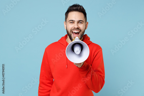 Young caucasian attractive happy bearded handsome leader man 20s wearing casual red orange hoodie screaming hot news shouting in megaphone loudly isolated on blue color background studio portrait