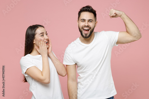 Young cheerful strong sporty fitness couple two friends man woman 20s in white basic blank print design t-shirts showing biceps muscles on hand isolated on pastel pink color background studio portrait