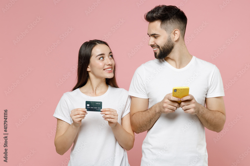 Young couple two friends man woman in white basic blank print design tshirts hold in hand using mobile cell phone credit bank card order home delivery in restaurant isolated on pastel pink background Young couple two friends man woman in white basic blank print design tshirts hold in hand using mobile cell phone credit bank card order home delivery in restaurant isolated on pastel pink background