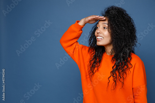 Smiling cheerful young african american woman wearing casual basic orange sweatshirt standing holding hand at forehead looking far away distance isolated on blue color background studio portrait.