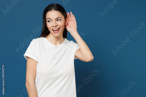 Young happy curious newsy nosy spanish smiling latin woman 20s wearing white casual basic t-shirt try to hear you overhear listening intently isolated on dark blue color background studio portrait. © ViDi Studio