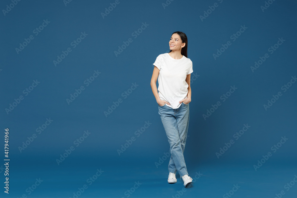 Full length of young smiling confident nice attractive beautiful latin woman 20s wearing white casual basic t-shirt keep hands in pockets look aside isolated on dark blue background studio portrait. Full length of young smiling confident nice attractive beautiful latin woman 20s wearing white casual basic t-shirt keep hands in pockets look aside isolated on dark blue background studio portrait.