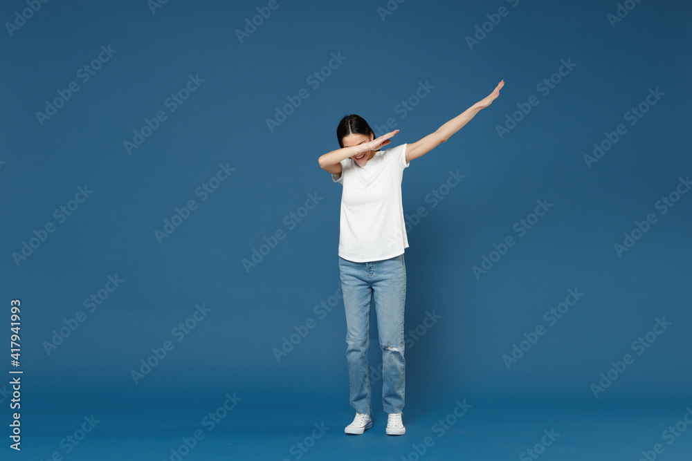 Full length young brunette woman 20s wearing white casual basic t-shirt doing dab hip hop dance hands move gesture youth sign hiding covering face isolated on dark blue background studio portrait. Full length young brunette woman 20s wearing white casual basic t-shirt doing dab hip hop dance hands move gesture youth sign hiding covering face isolated on dark blue background studio portrait.