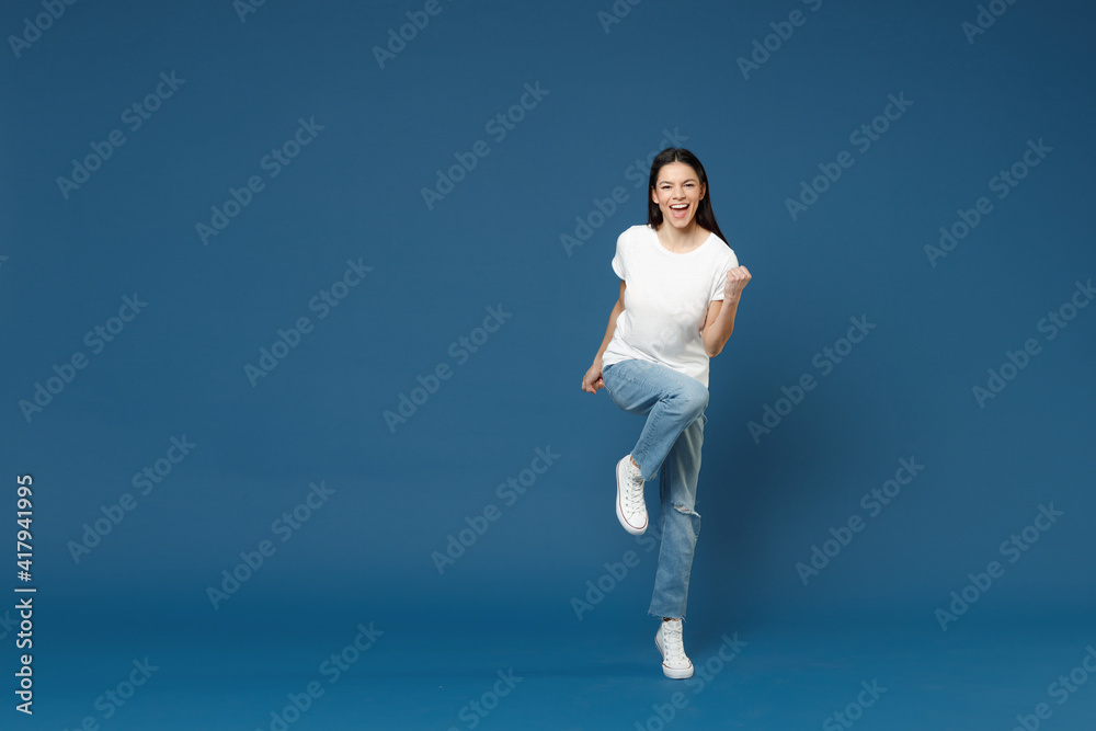 Full length young happy excited overjoyed latin woman 20s in white casual basic t-shirt do winner gesture clench fist raised up leg look camera isolated on dark blue color background studio portrait.