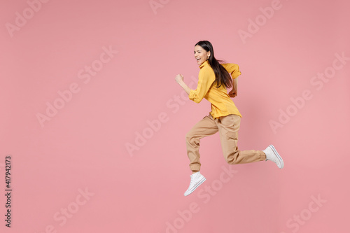 Full length side view of young smiling brunette excited overjoyed attractive latin woman 20s in yellow shirt running fast jumping high hurrying isolated on pastel pink background studio portrait