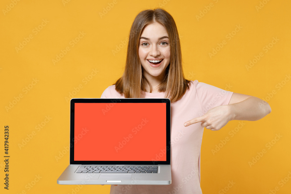 Naklejka premium Young surprised freelancer woman 20s wearing casual basic pastel pink t-shirt point index finger on laptop pc computer with blank screen workspace area isolated on yellow background studio portrait
