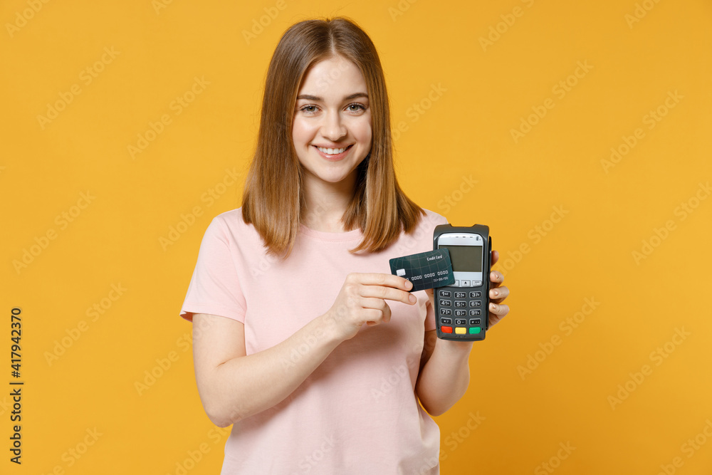 Young woman in basic pastel pink t-shirt hold wireless modern bank payment terminal to process and acquire bank credit card payments show ok okay gesture isolated on yellow background studio portrait.