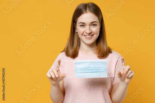 Young smiling happy woman 20s wearing basic pastel pink t-shirt hold sterile face mask to safe from coronavirus virus covid-19 during pandemic quarantine isolated on yellow background studio portrait