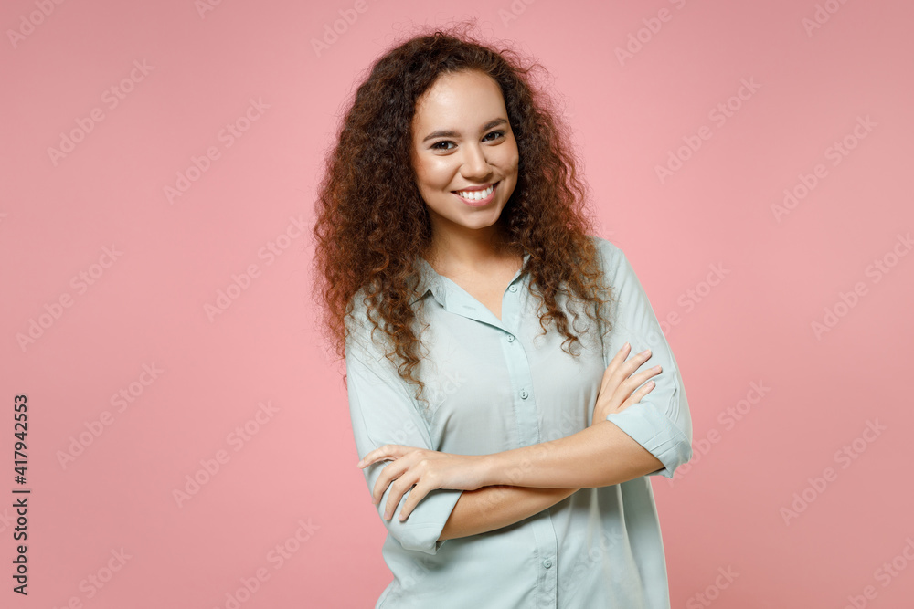 Young black african american happy confident smiling positive curly woman 20s wearing blue shirt hold hands crossed folded isolated on pastel pink background studio portrait. People lifestyle concept. Young black african american happy confident smiling positive curly woman 20s wearing blue shirt hold hands crossed folded isolated on pastel pink background studio portrait. People lifestyle concept.