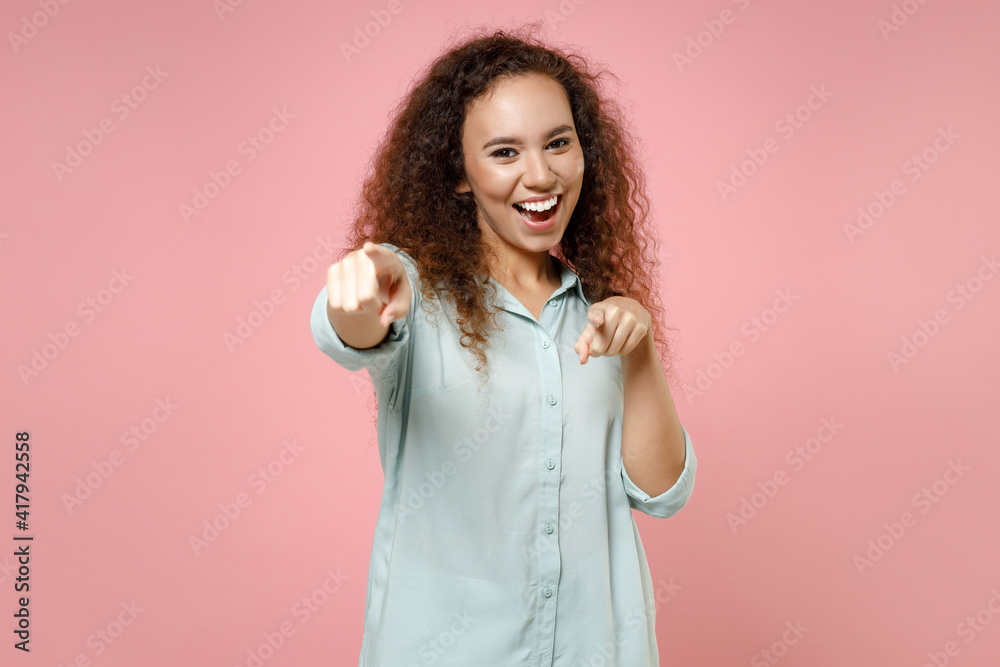 Young black african american fun happy leader friendly positive curly woman 20s in blue shirt point index finger camera on you motivating encourage isolated on pastel pink background studio portrait