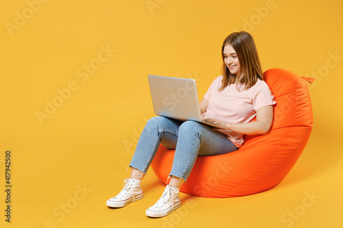 Full length of young student woman 20s in basic pastel pink t-shirt, jeans sitting in orange bean bag chair holding laptop pc computer browsing internet in lounge zone isolated on yellow background.