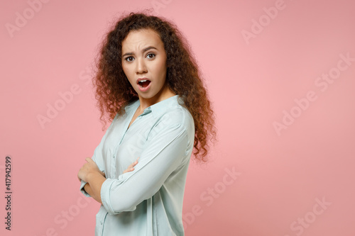 Young black african american indignant arrogant shocked curly woman 20s wearing casual blue shirt holding hands crossed folded looking camera isolated on pastel pink color background studio portrait.
