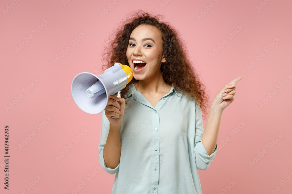 Young black african american surprised excited cute rich curly woman 20s in blue shirt screaming in megaphone point index finger aside on workspace isolated on pastel pink background studio portrait. Young black african american surprised excited cute rich curly woman 20s in blue shirt screaming in megaphone point index finger aside on workspace isolated on pastel pink background studio portrait.