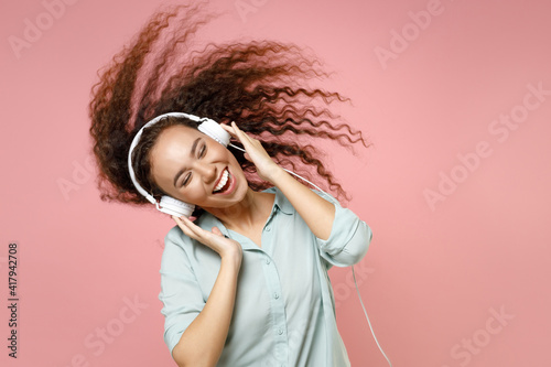 Young black african american fun student happy smiling positive friendly curly woman in blue casual shirt wear headphones dancing fluttering hair isolated on pastel pink background studio portrait photo
