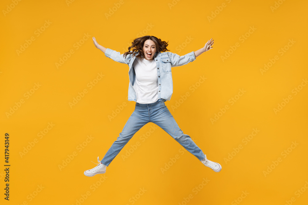 Full length of young overjoyed excited fun expressive student happy woman 20s wearing denim shirt white t-shirt with outstretched hands legs jump high isolated on yellow background studio portrait