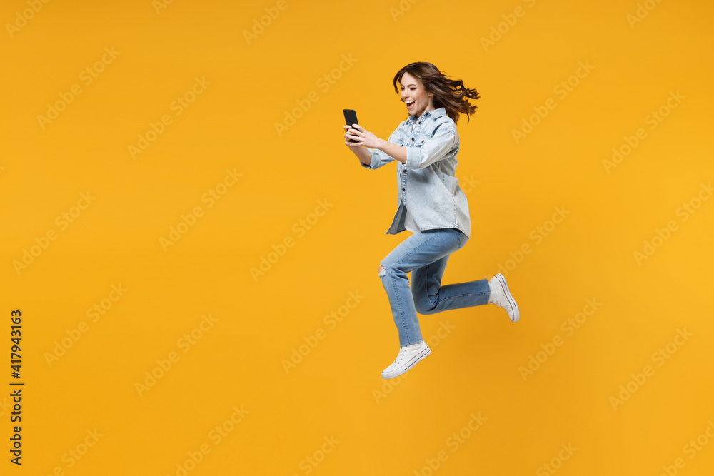 Full length of young fun sporty student woman 20s wear casual stylish denim shirt white t-shirt run jump high hold mobile cell phone use fast internet isolated on yellow background studio portrait