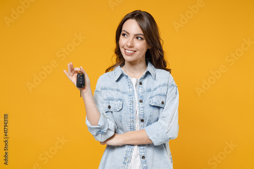 Young smiling dreamful pensive wistful happy rich brunette woman 20s wearing stylish denim shirt white t-shirt holding in hands car keys look aside isolated on yellow color background studio portrait.