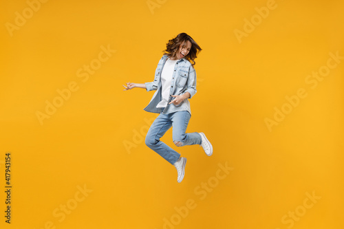 Full length of young overjoyed excited fun expressive student happy woman 20s wearing casual denim shirt white t-shirt playing guitar jump high isolated on yellow color background studio portrait